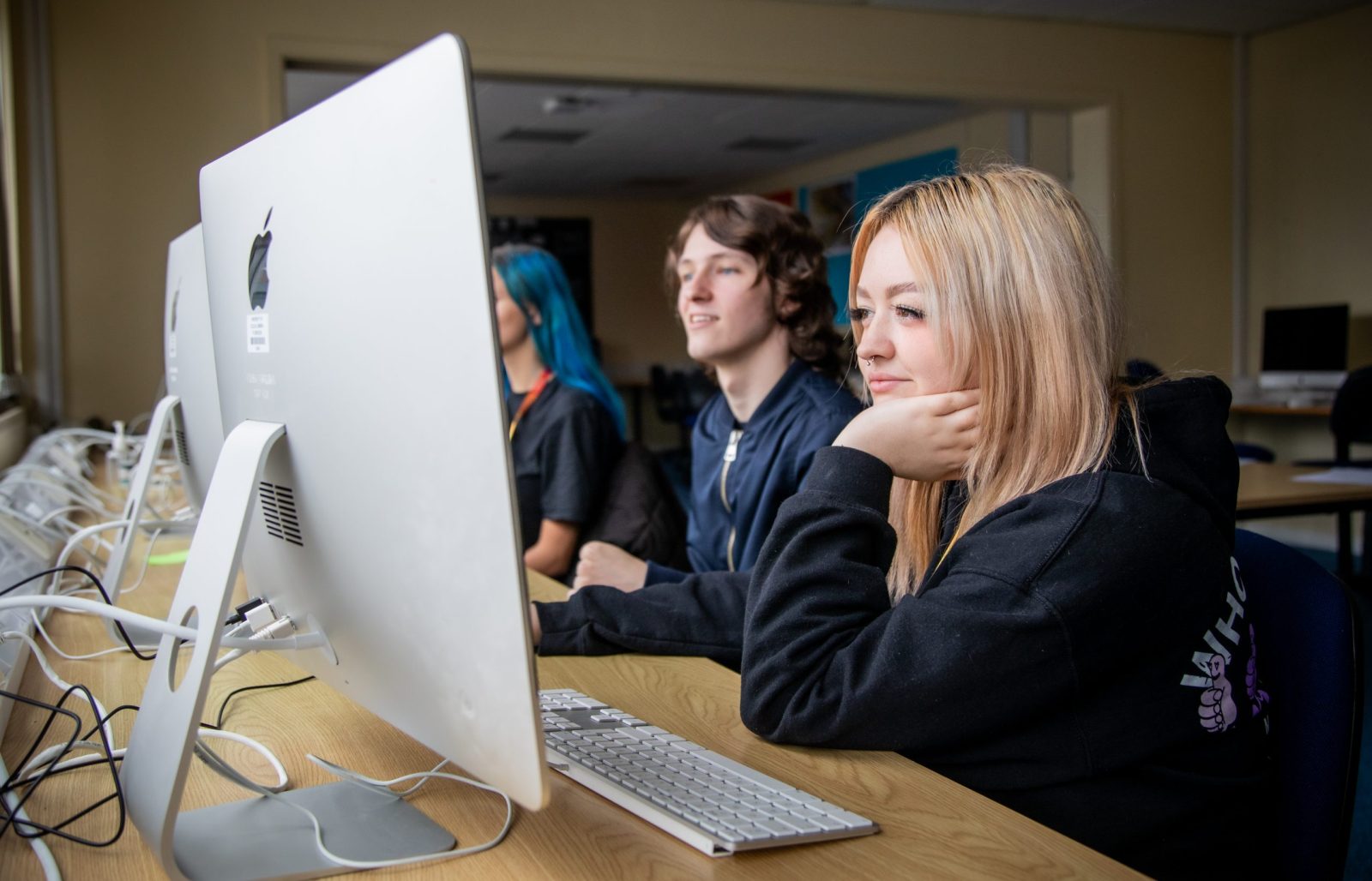 Myfyrwyr yn eistedd wrth fyrddau gan ddefnyddio cyfrifiaduron iMac mewn ystafell ddosbarth, yn canolbwyntio ar eu sgriniau, gyda un myfyriwr yn gorffwys ei gên ar ei llaw.