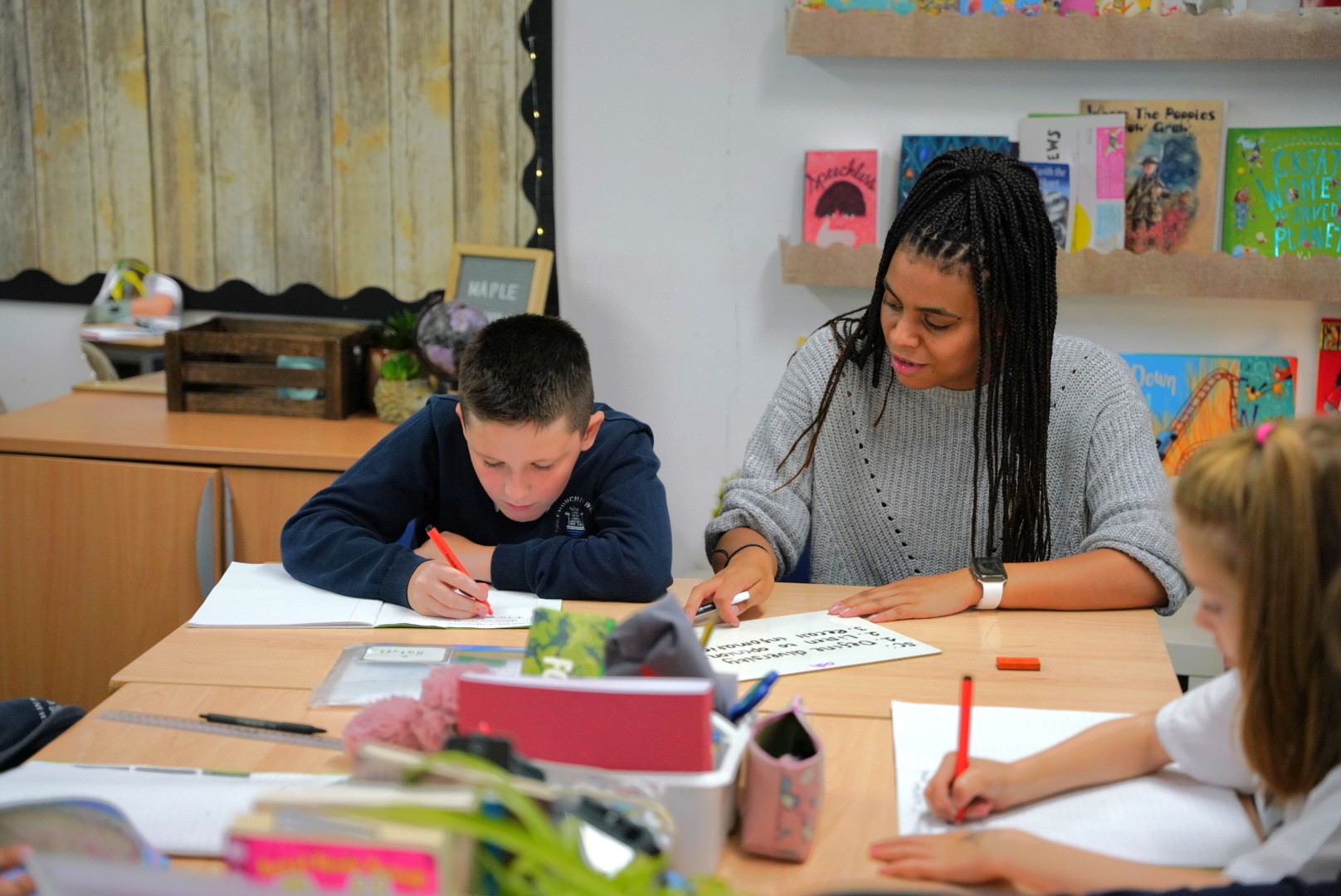 A teacher is sitting at a desk, guiding a young student in a classroom setting with books and educational materials around.
