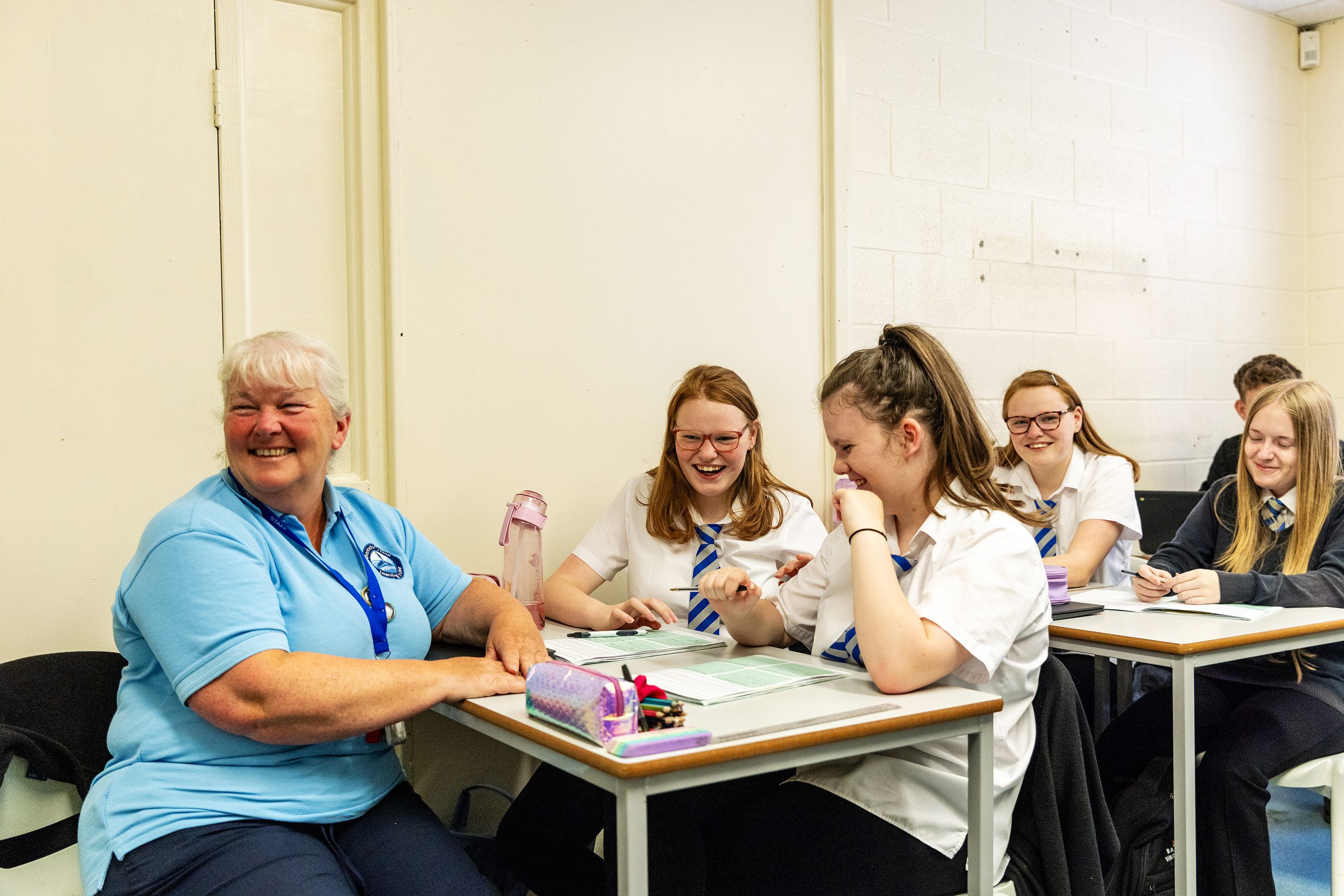 A teacher with white hair and a blue shirt is sitting at a desk, laughing with a group of students in school uniforms. The students are also laughing and appear to be enjoying the interaction.