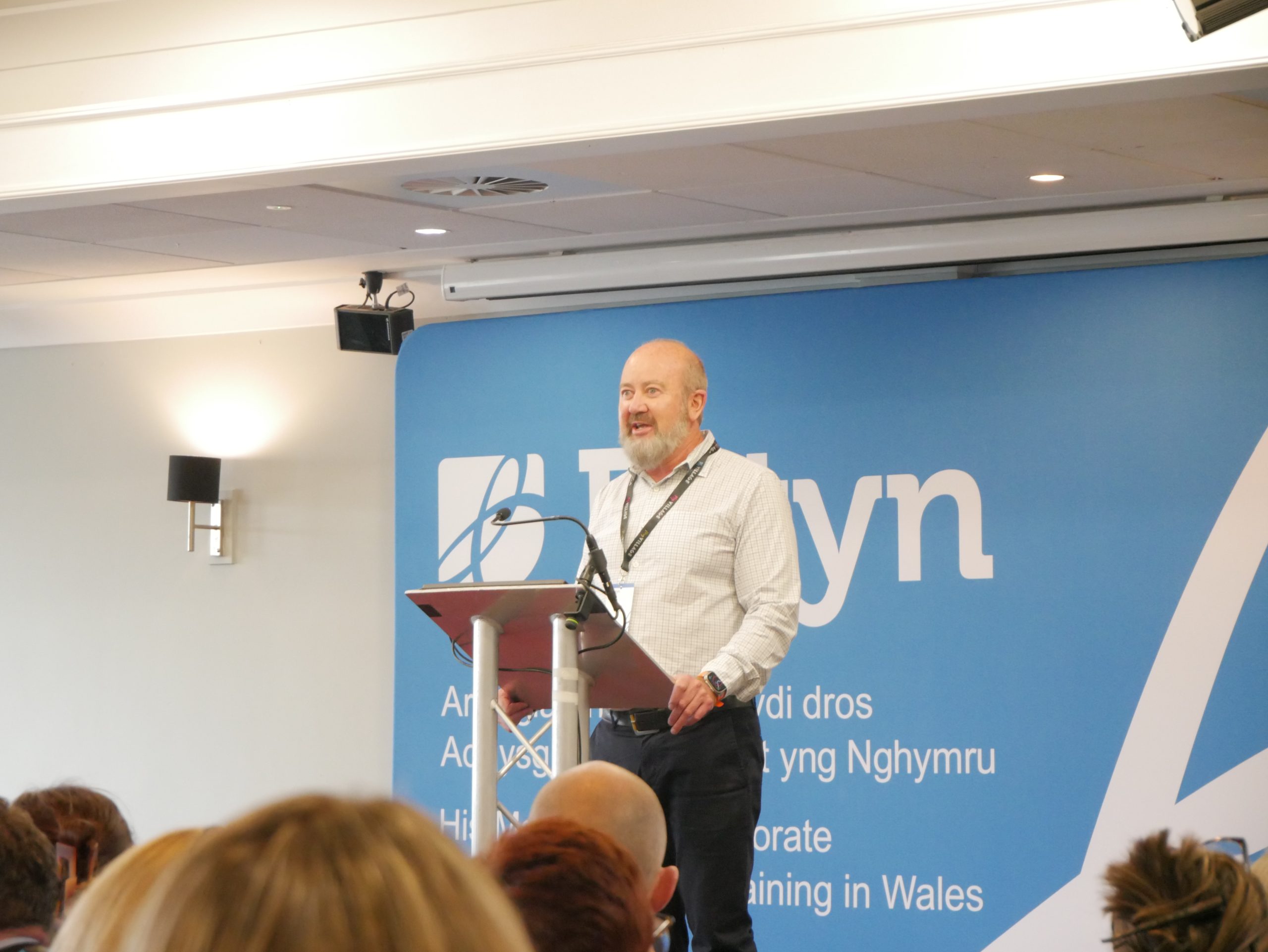 Dyn yn traddodi araith wrth bulpud o flaen cefndir glas gyda'r geiriau "Estyn" ac eraill, gan annerch cynulleidfa mewn digwyddiad.