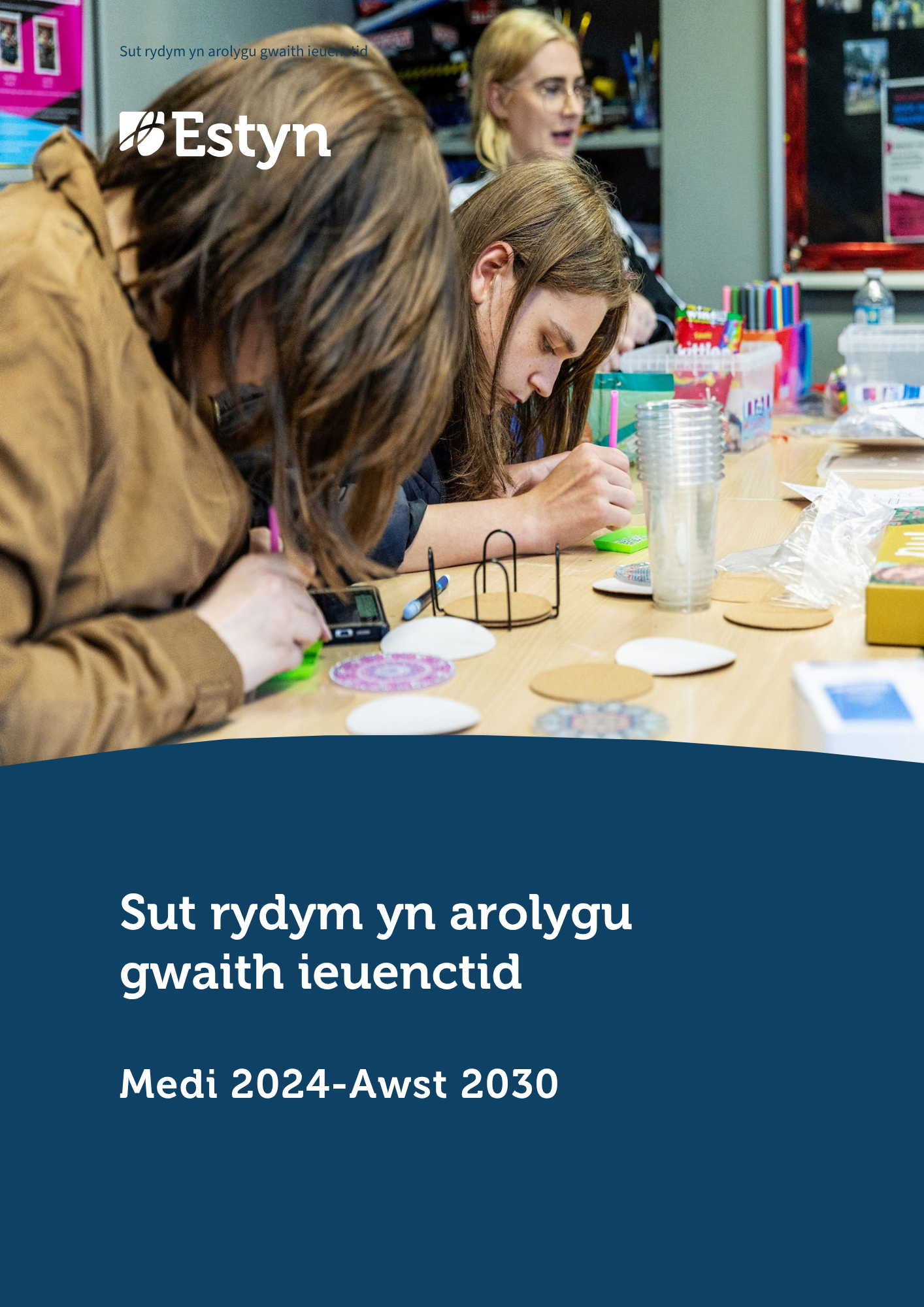 Hyfforddwr a dysgwr yn gweithio gyda’i gilydd mewn gweithdy, yn adolygu mesuriadau a chynlluniau wrth fainc gwaith coed wedi’i hamgylchynu gan offer a deunyddiau.