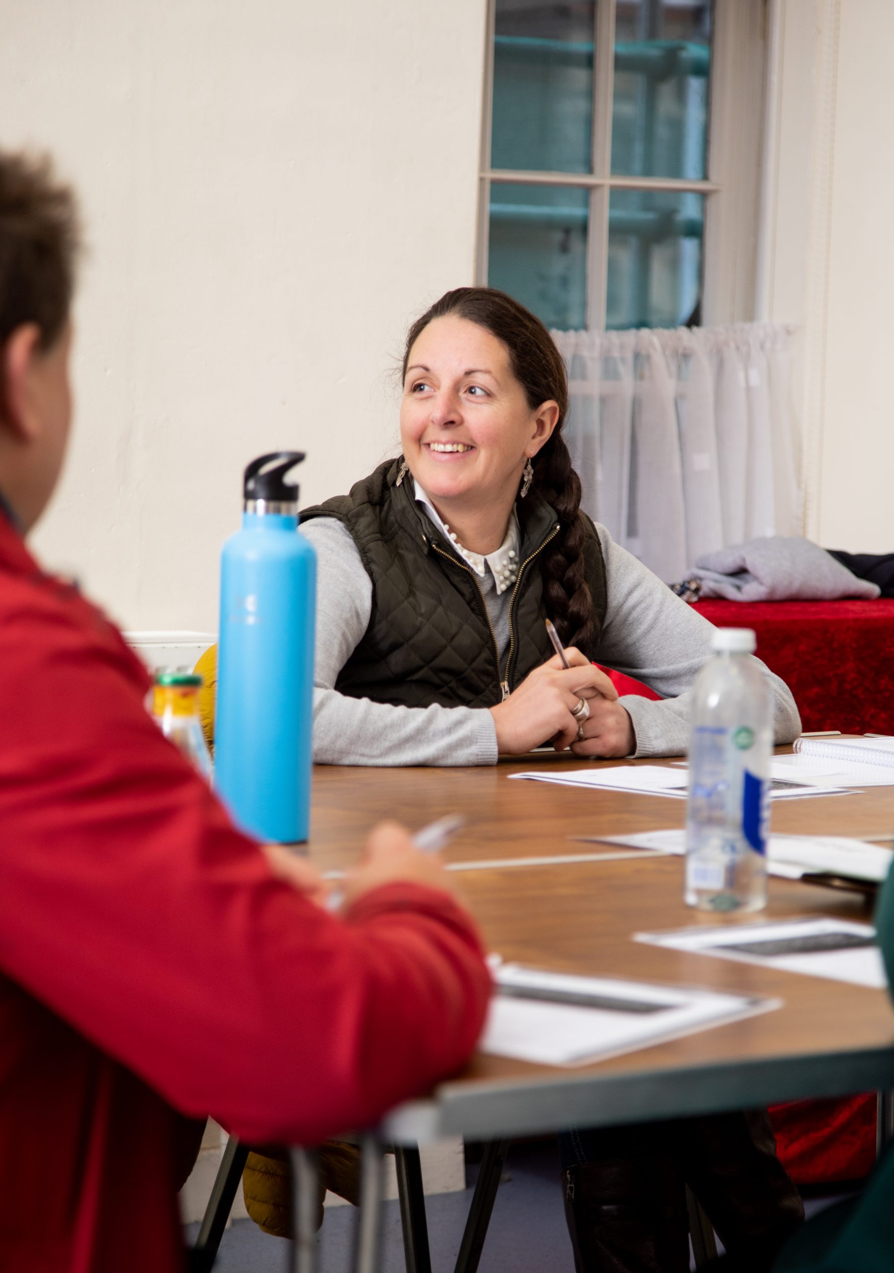 A woman in a vest sitting at a table, smiling and looking towards someone across the table during a meeting.