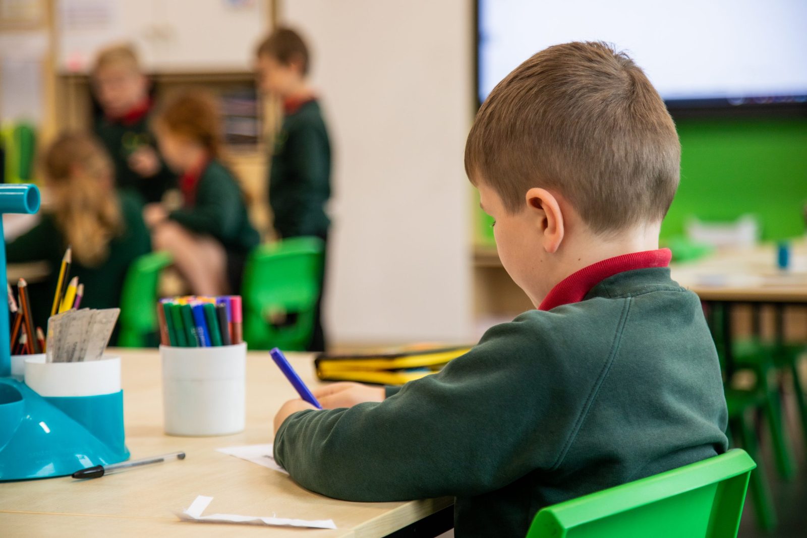 A boy sitting at a table, focused on writing or drawing, with other children in the background.
