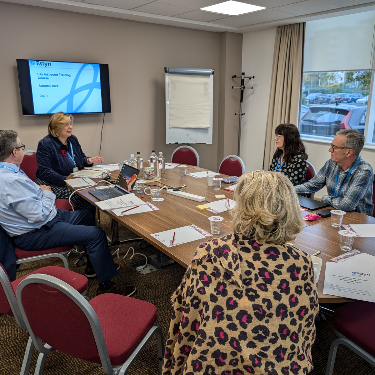 A group of professionals sitting around a conference table discussing, with a presentation labeled "Estyn" on a screen in the background.