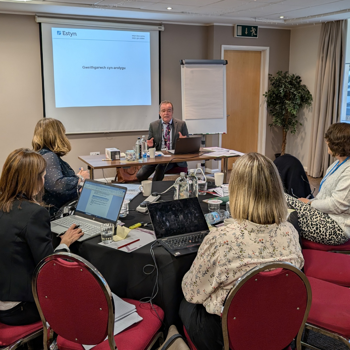 A professional meeting with several participants seated around tables in a conference room. The focus is on an individual at the front presenting with a screen behind them displaying the logo of Estyn. Participants are watching and using laptops and notepads.