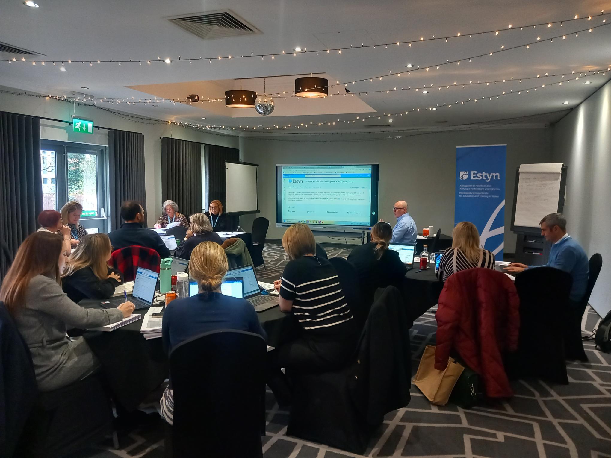 Attendees at a corporate workshop sitting at tables in a conference room, facing a presenter and a large screen displaying a PowerPoint presentation. A banner reading 'Estyn' is visible in the background.