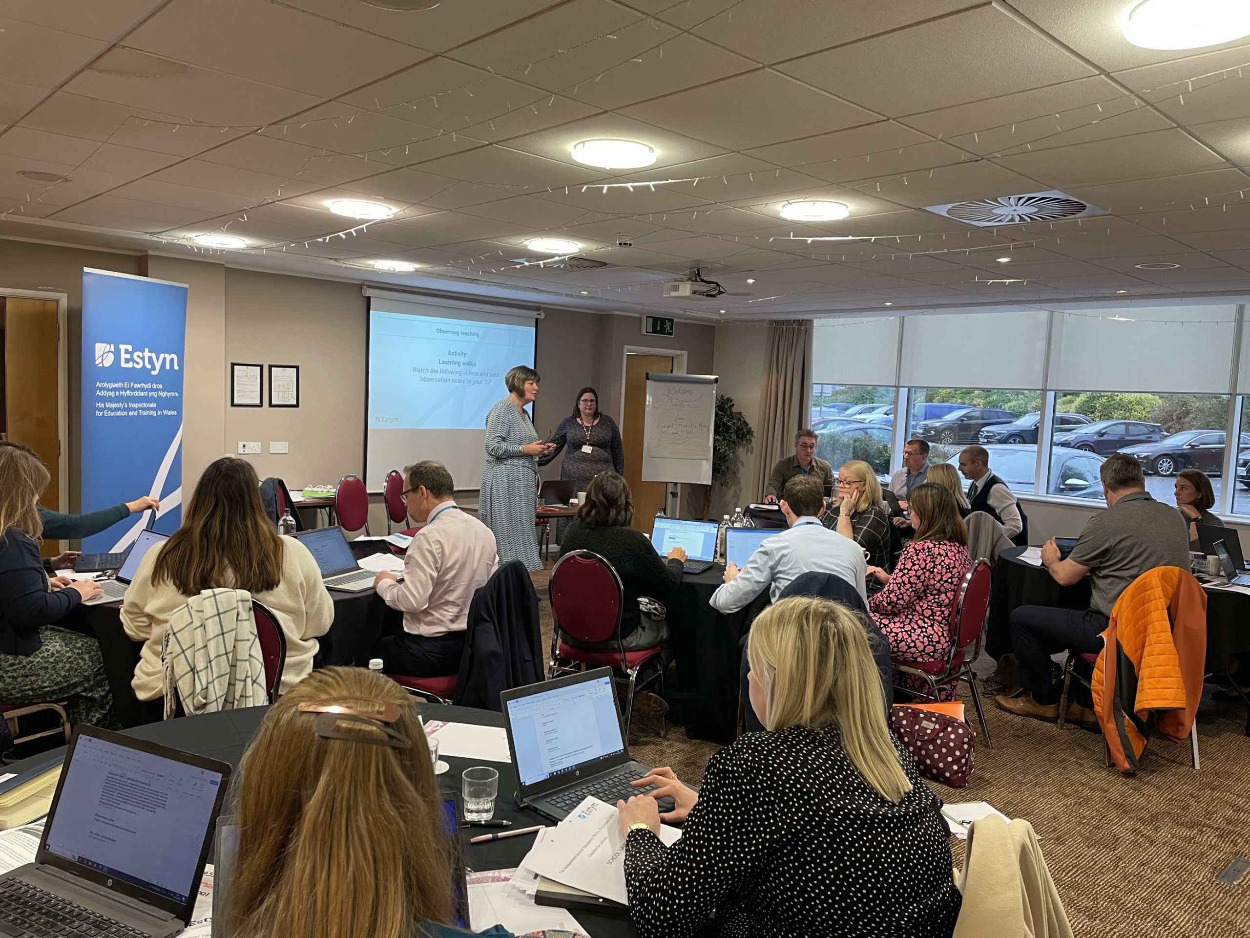 Estyn workshop with participants seated at tables, engaging in discussions and taking notes; two presenters speaking near a flip chart at the front of the room.