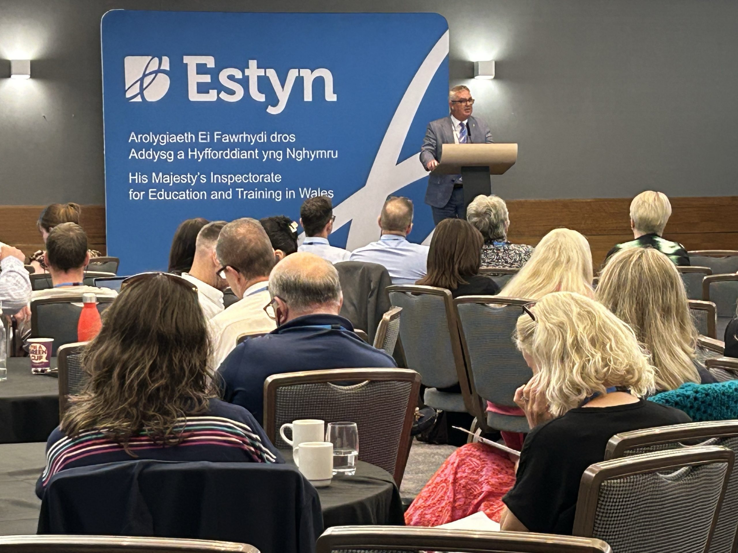 A person presenting at a conference in front of a banner for Estyn, His Majesty's Inspectorate for Education and Training in Wales. The audience appears engaged and attentive.