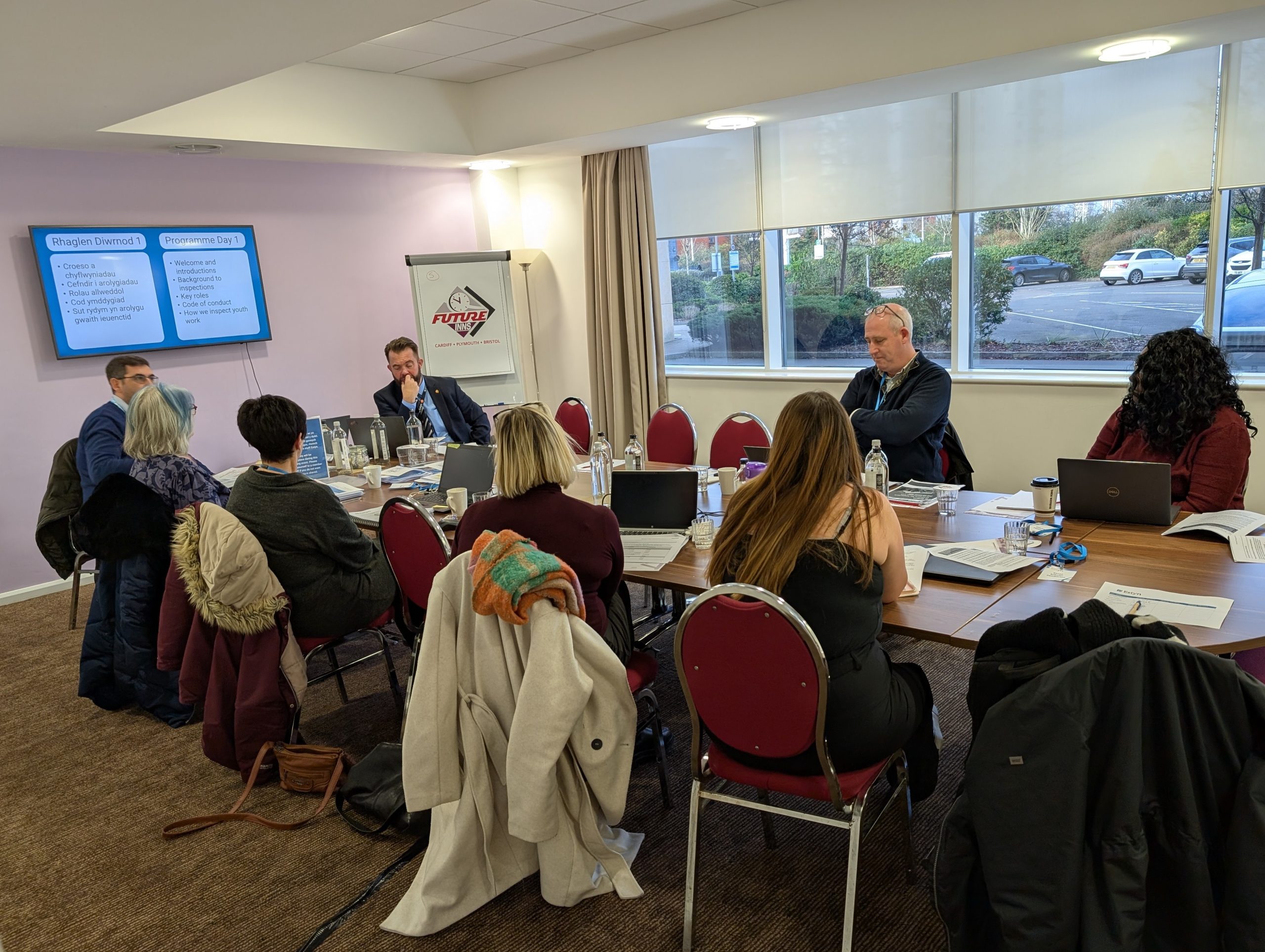 Group of professionals seated around a table in a meeting room, discussing information displayed on a projector screen with the logo of Premier Inn visible.