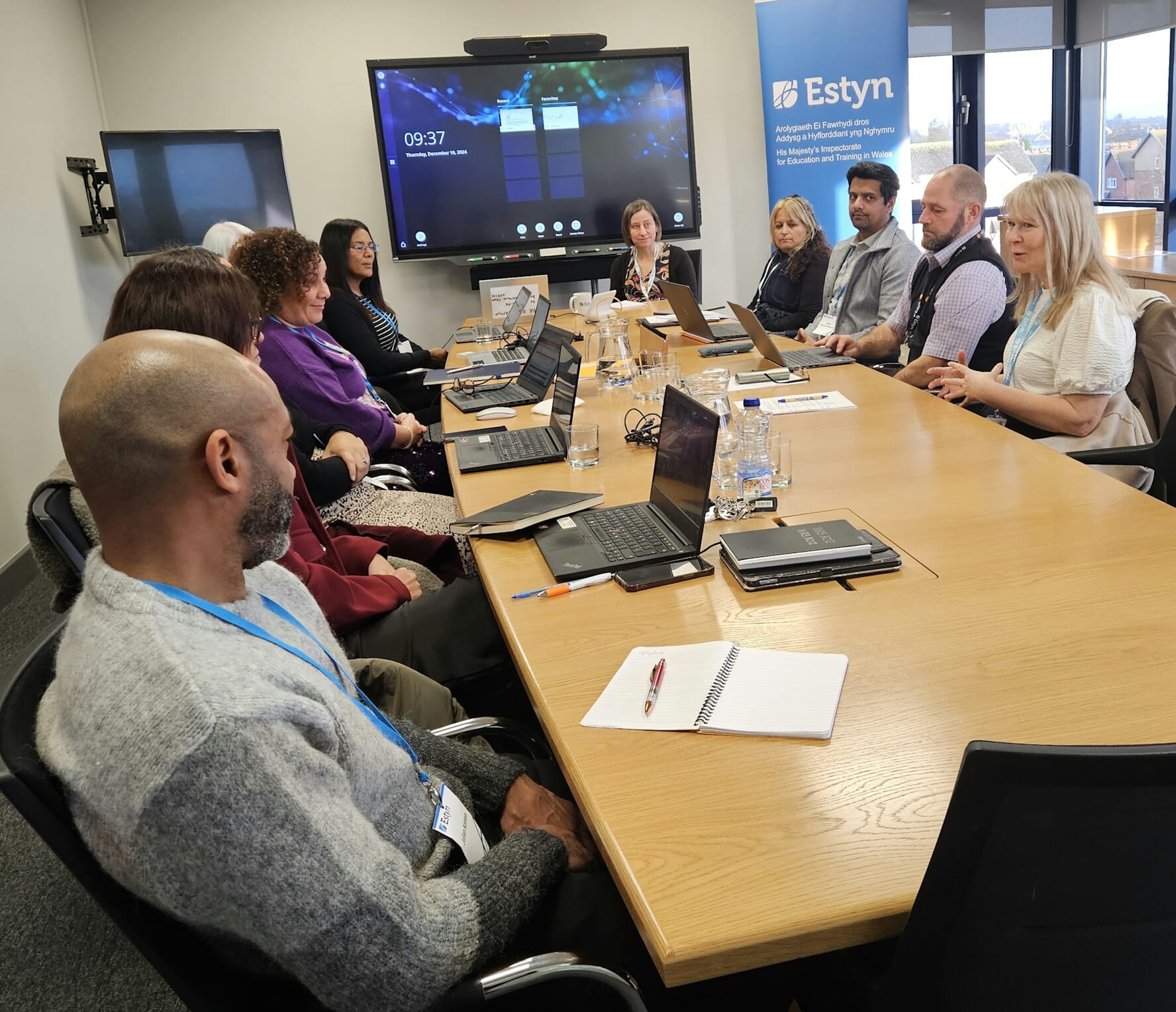 Group of professionals sitting around a conference table with laptops, engaged in a meeting at an Estyn office.