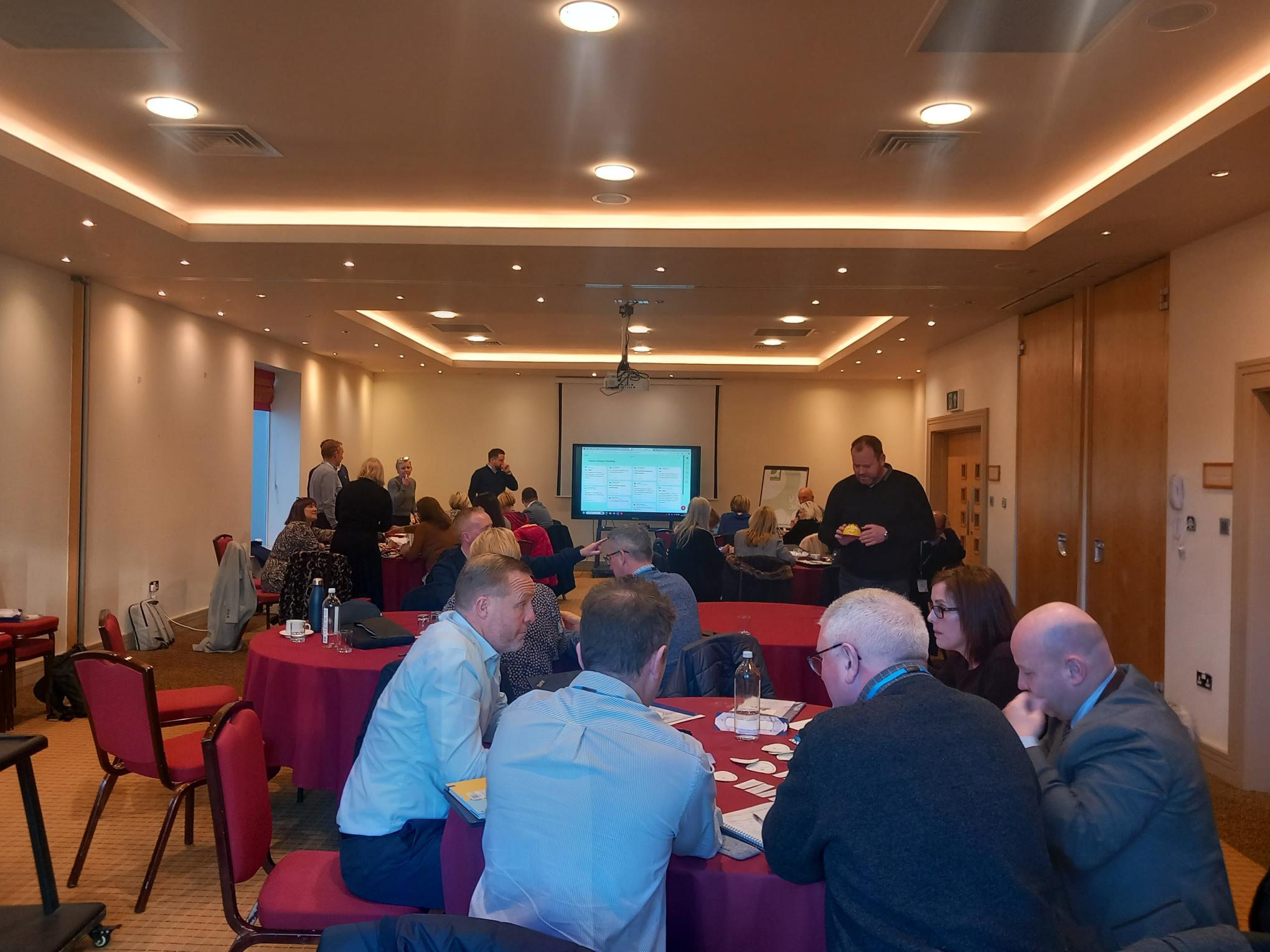 A business workshop in a conference room with attendees seated around tables, engaging in discussions. A presentation screen displays information visible in the background.