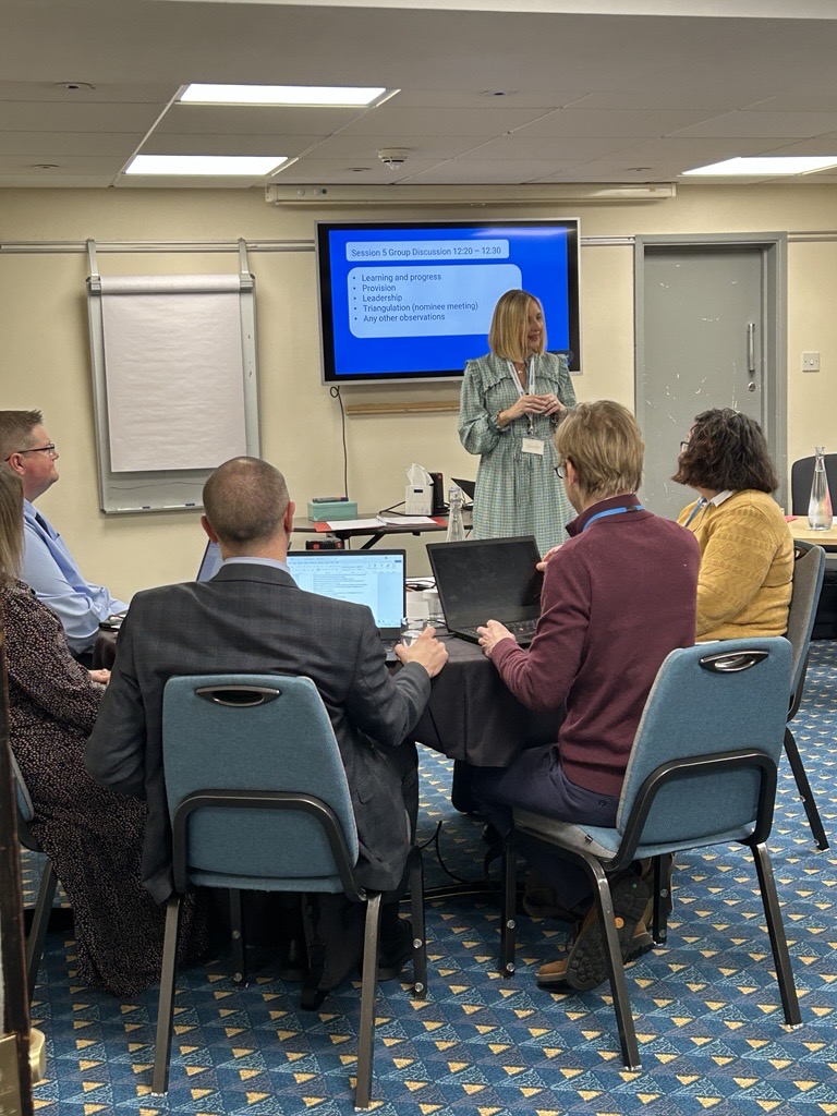 A presenter standing in front of a screen displaying a PowerPoint presentation at a professional seminar, addressing an audience of seated adults