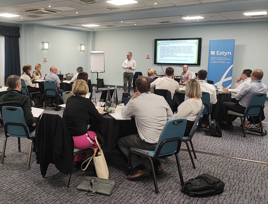 Professional meeting in progress at an Estyn event, with attendees seated around tables listening to a presenter standing near a screen displaying a PowerPoint presentation.