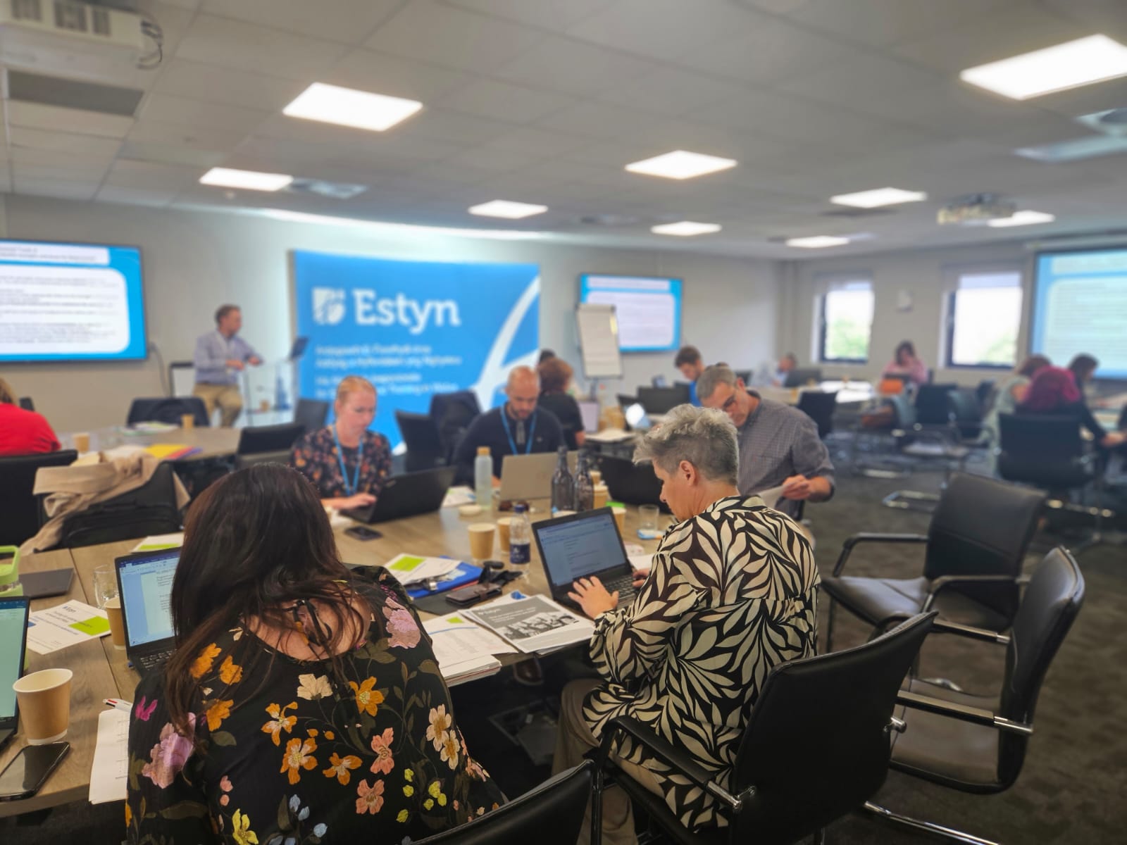 Group of professionals engaged in a workshop at Estyn, focusing on discussions and laptops, in a meeting room with informational banners in the background.