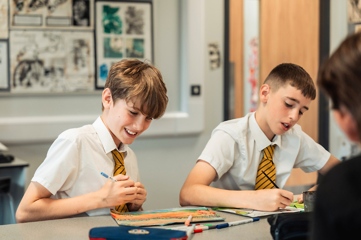 Mae dau fyfyriwr mewn gwisgoedd ysgol gyda thei streipiog yn eistedd wrth ddesg, yn sgwrsio ac yn gweithio ar brosiect celf mewn ystafell ddosbarth wedi'i haddurno â gweithiau celf amrywiol.