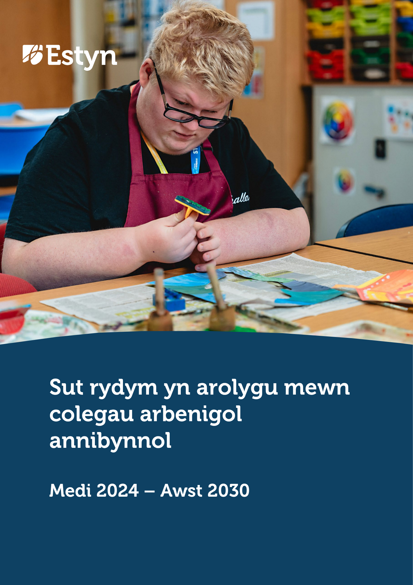 Myfyriwr yn gwisgo sbectol a ffedog yn canolbwyntio ar beintio prosiect crefft lliwgar mewn ystafell ddosbarth.