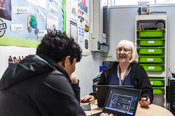 Athrawes yn gwenu yn eistedd ar fwrdd dosbarth gyferbyn â disgybl.