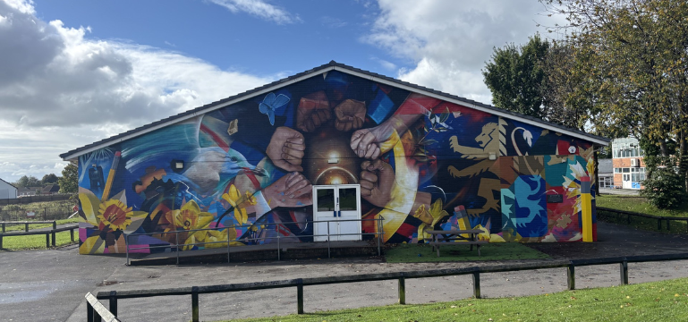 Mae'r ddelwedd yn dangos murlun lliwgar ar ochr adeilad, yn darlunio dwylo amrywiol o wahanol arlliwiau croen wedi'u casglu o amgylch pêl sy'n tywynnu. O amgylch y dwylo mae elfennau artistig bywiog, gan gynnwys blodau, siapiau haniaethol, a llew. Mae'r olygfa wedi'i gosod yn yr awyr agored o dan awyr rhannol gymylog.