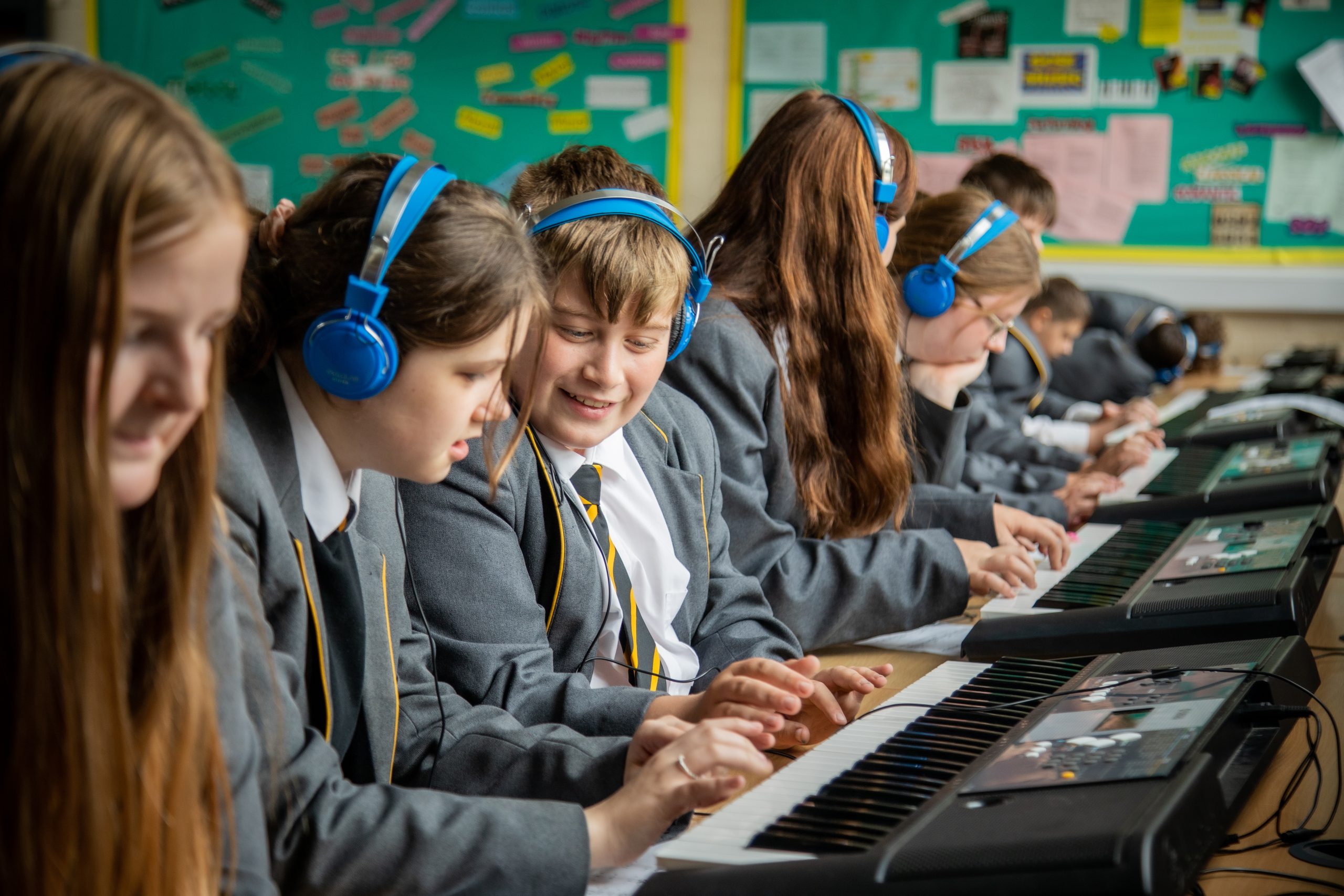 Disgyblion mewn gwisg ysgol yn eistedd ochr yn ochr wrth fysellfyrddau electronig, yn gwisgo clustffonau glas ac yn canolbwyntio ac yn cydweithio yn ystod gwers gerddoriaeth mewn ystafell ddosbarth.