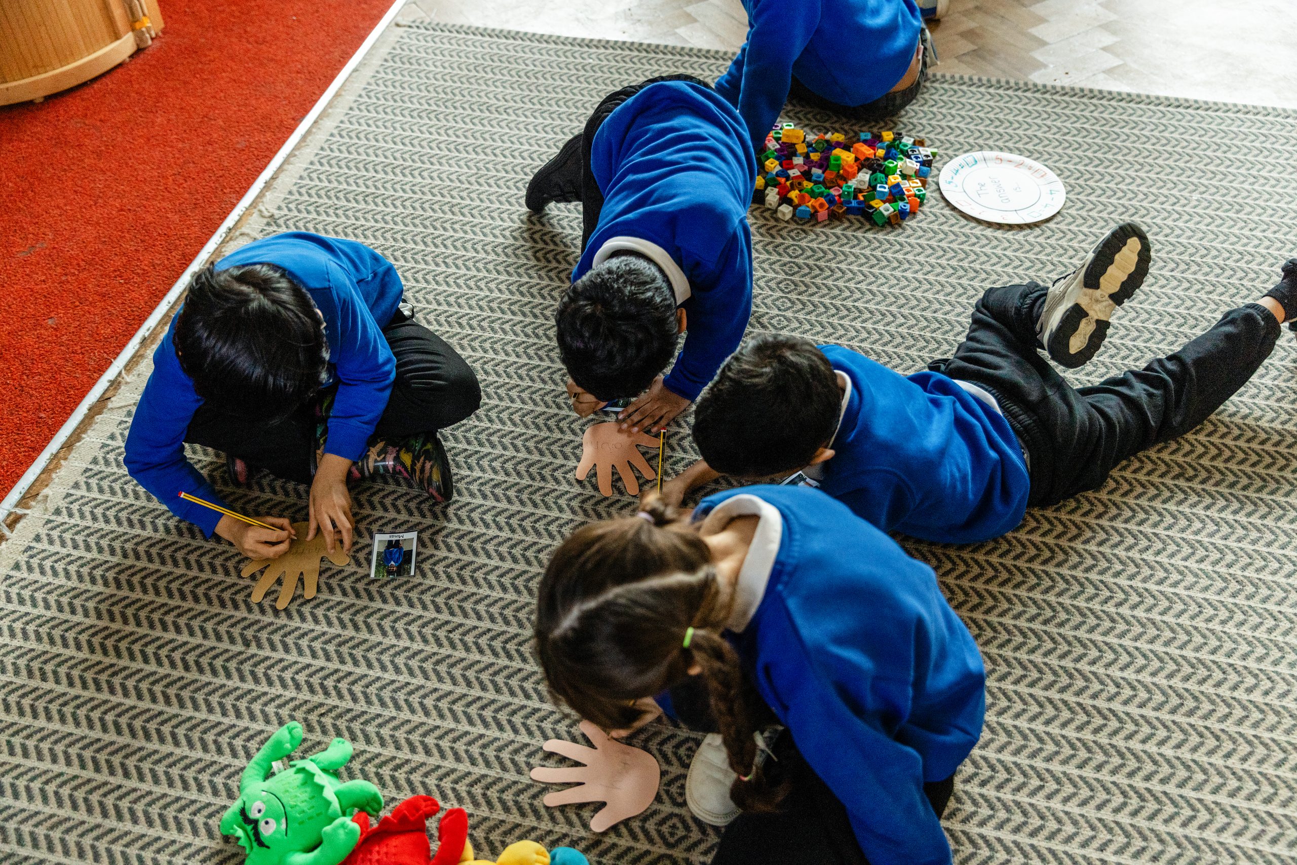 A group of primary school pupils in blue school uniforms sit and kneel on a classroom carpet, working together on a hands-on learning activity. They are writing on paper hand cut-outs with pencils, while colourful counting cubes, a teaching clock and soft toys are nearby.